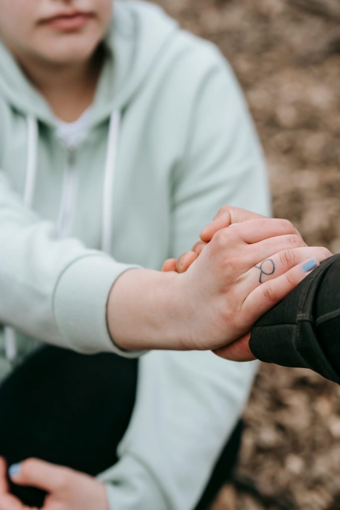 From above of crop anonymous overweight woman sitting on ground and holding hand of unrecognizable person in daytime